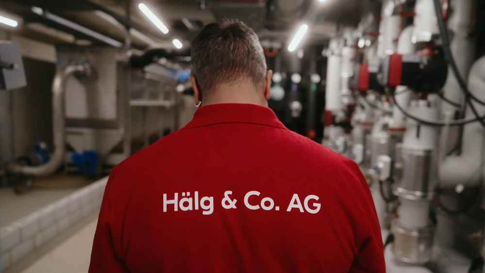 Rückenansicht eines Mitarbeiters im roten Poloshirt mit Aufdruck „Hälg & Co. AG“ in einem Technikraum.