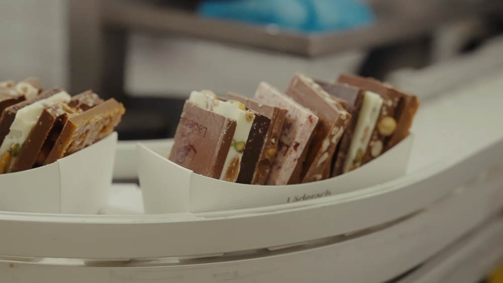 Läderach chocolatier suisse - Schokaldenproduktion, mehrere Täfelchen auf Laufband.